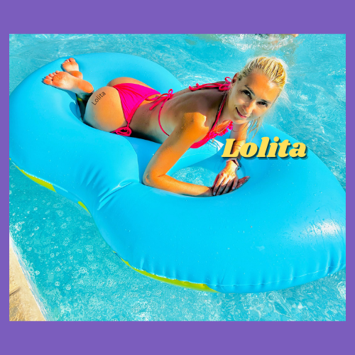Lolita of IndieCamFans is wearing a pink pathing suit, her blonde hair is tied back and she is relaxing on her belly on a blue floaty which is resting in a pool.